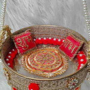 Op de afbeelding: Een ronde goudkleurige decoratieve mand met ingewikkelde metalen gaaswanden, een centrale ronde mat met gouden borduursels en twee kleine rode vierkante kussentjes met gouden Hindi-schrift. De mand is versierd met rode pompons en witte parels.