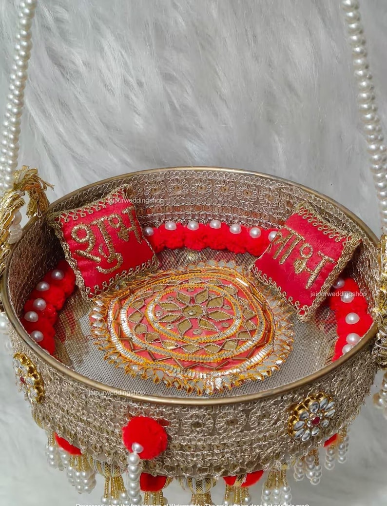 Op de afbeelding: Een ronde goudkleurige decoratieve mand met ingewikkelde metalen gaaswanden, een centrale ronde mat met gouden borduursels en twee kleine rode vierkante kussentjes met gouden Hindi-schrift. De mand is versierd met rode pompons en witte parels.