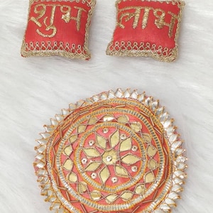 Op de afbeelding: Een rond gewatteerd bed en twee vierkante kussens. Het ronde bed heeft een roze middenstuk met gouden bloemenborduursel, zilveren metallic afwerking en een geschulpte rand. De twee rode kussens hebben gouden borduursels en gouden randen.