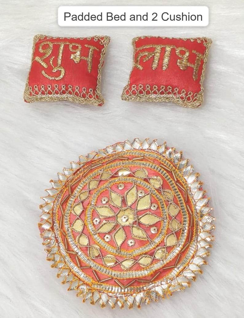 Op de afbeelding: Een rond gewatteerd bed en twee vierkante kussens. Het ronde bed heeft een roze middenstuk met gouden bloemenborduursel, zilveren metallic afwerking en een geschulpte rand. De twee rode kussens hebben gouden borduursels en gouden randen.