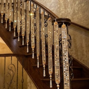 May include: A series of long, vertical decorative fabric strips draped over a wooden staircase railing. Each strip features a gold-toned border, a central panel of small circular mirrors, and a dangling white beaded tassel at the bottom end.