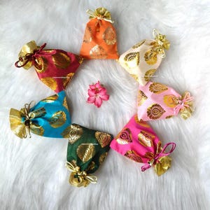 Könnte beinhalten: Eine Anordnung von farbigen Stoffbeuteln in einem Kreis, verziert mit goldenen Mustern und Kordelzügen. Die Beutel sind in Orange, Rot, Blau, Grün, Rosa und Creme gehalten, mit einer rosa Blume in der Mitte.
