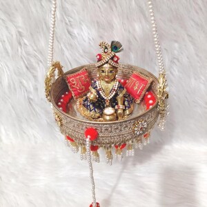 Op de afbeelding: Een gouden ronde schommel met een sierlijk filigraanpatroon, opgehangen aan parelkettingen. Binnenin zit een klein godenbeeldje in een blauw met gouden outfit, geflankeerd door twee rode kussentjes met gouden schrift en rode pompons.