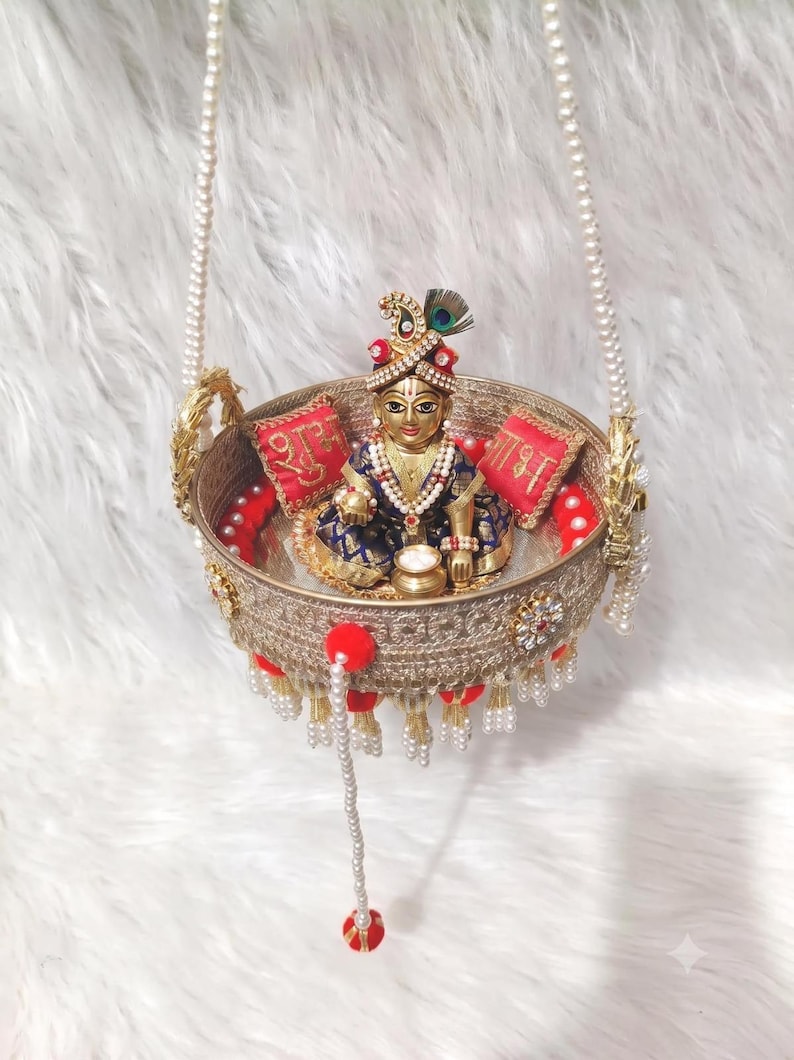 Op de afbeelding: Een gouden ronde schommel met een sierlijk filigraanpatroon, opgehangen aan parelkettingen. Binnenin zit een klein godenbeeldje in een blauw met gouden outfit, geflankeerd door twee rode kussentjes met gouden schrift en rode pompons.