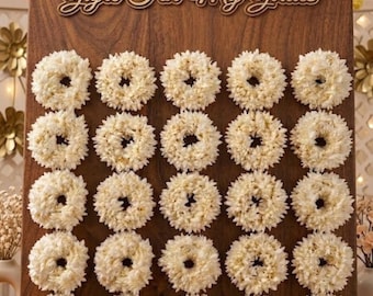 May include: A rectangular wooden display board featuring the text "Gajre For My Girlies" at the top. The board is arranged with twenty-five circular white floral wreaths in a five-by-five grid pattern, each made of small, densely packed white petals.
