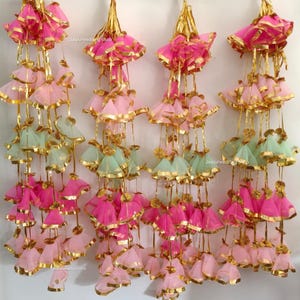 May include: A collection of hanging decorative ornaments in shades of pink, light pink, and mint green, with gold accents. The ornaments are made of sheer fabric and are suspended from gold-colored cords. The text "Jaipurwedding shop" is visible.