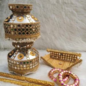 May include: A decorative silver-toned metal pot adorned with gold trim, circular mirrors, and white beaded fringe. Accompanying the pot is a rectangular gold-trimmed tray with mirror accents, two pink and gold striped bangles, and two gold sticks.