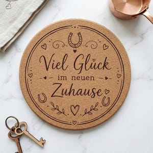 Könnte beinhalten: Ein runder Korkuntersetzer mit der lasergravierten Aufschrift Viel Glück im neuen Zuhause, umgeben von dekorativen Hufeisen-, Herz- und Blattmotiven. Der Untersetzer wird von zwei Metallschlüsseln und einem kupferfarbenen kleinen Topf begleitet.