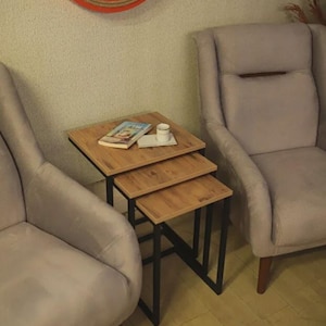 May include: A set of three nesting tables with a light wood top and black metal legs. A book and a small white cup and saucer sit on the top table. Two gray armchairs are on either side of the tables.