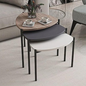 May include: A set of three nesting tables with a modern design. The top table is wood-toned, the middle is dark gray, and the bottom is white. Each table has black metal legs. Two cups and saucers sit on the top table.