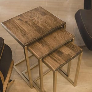Op de afbeelding: Set van drie nesttafels met vierkante houten bladen en goudkleurige metalen frames. Het hout heeft een warme, bruine tint met zichtbare nerf. De tafels zijn in aflopende grootte gerangschikt, waardoor een gelaagd effect ontstaat. Modern design.
