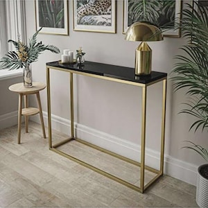 Könnte beinhalten: Eine moderne Konsole mit schwarzer Oberfläche und goldfarbenem Metallrahmen. Auf dem Tisch stehen eine goldfarbene Lampe, Kerzen und eine Vase. Gerahmte Kunstwerke hängen an der Wand hinter dem Tisch.