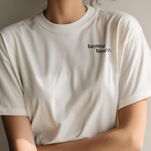 Könnte beinhalten: Cremefarbenes T-Shirt mit dem schwarzen Aufdruck "beyond binary.". Das Shirt hat kurze Ärmel und einen Rundhalsausschnitt. Die Person trägt eine blaue Jeans.