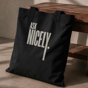Op de afbeelding: Een zwarte tote bag met de tekst ASK NICELY gedrukt in een groot, lichtgekleurd schreefloos lettertype. De tas heeft twee stoffen handvatten aan de bovenkant en een rechthoekige vorm. Het ontwerp is gecentreerd op het zwarte oppervlak.