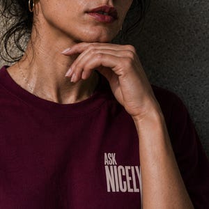 Puede incluir: Una camiseta de color burdeos oscuro con el texto "ASK NICELY" impreso en una fuente sans-serif de color beige claro en el pecho. La prenda tiene un cuello redondo clásico y mangas cortas, ofreciendo un diseño sencillo y directo.