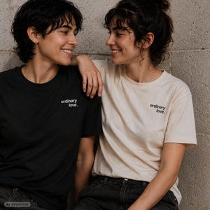 Könnte beinhalten: Ein schwarzes Kurzarm-T-Shirt mit dem weißen Schriftzug 'ordinary love.' auf der Brust und ein cremefarbenes Kurzarm-T-Shirt mit dem schwarzen Schriftzug 'ordinary love.' auf der Brust, beide mit einem klassischen Rundhalsausschnitt.