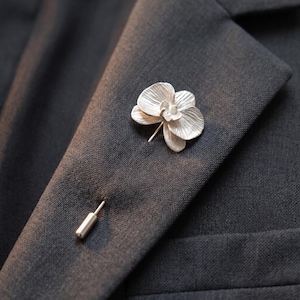 May include: A silver-toned orchid flower lapel pin featuring textured petals and a central column, attached to a long, slender metal pin stem with a cylindrical end cap, secured through the fabric of a dark grey textured suit jacket lapel.