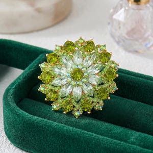 May include: A floral brooch with a gold-tone base, featuring a central cluster of clear, faceted stones surrounded by layers of green and chartreuse crystals. The brooch is set against a dark green velvet surface.