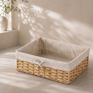 May include: A rectangular woven seagrass storage basket featuring a removable beige fabric liner. The liner is decorated with a repeating pattern of small white daisy flowers with yellow centers and includes a small fabric bow on the front side.