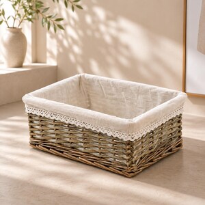 May include: A rectangular woven wicker basket featuring a natural brown finish. The interior is lined with a light beige fabric that folds over the top rim, accented by a delicate white lace trim along the edge of the fabric lining.