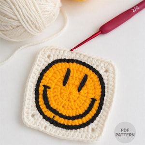 May include: A crocheted square featuring a yellow smiley face with black details, set against a white background. A ball of cream yarn and a red crochet hook are visible. The image also includes the text "PDF PATTERN" in the corner.