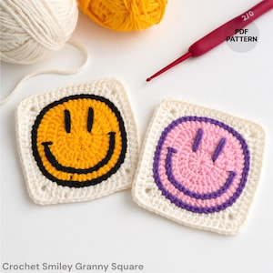 May include: Two crocheted granny squares with smiley faces. One square has a yellow smiley face with a black outline, and the other has a pink smiley face with a purple outline. The squares are on a white surface, with yarn and a crochet hook.