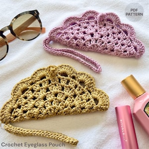 May include: Two handmade crochet eyeglass pouches in gold and lavender, with a scalloped edge design. The pouches have a wrist strap and are shown with sunglasses and beauty products. The text "PDF PATTERN" is visible.