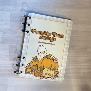 May include: A spiral-bound notebook with a cream and white checkered border. The cover features the text "Pumpkin Patch Savings" and "North Country Cash Queen" above an illustration of a white duck surrounded by orange pumpkins and a small lantern.