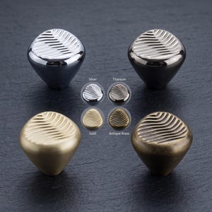 May include: Four cabinet knobs with a conical base and a rounded top featuring a textured, wavy, leaf-like pattern. The finishes include polished silver, dark titanium, matte gold, and antique brass, with labels for each colour variant shown.