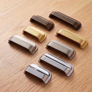 May include: A collection of eight metal drawer pulls arranged in two columns, featuring a ribbed, arched design. The pulls are available in four distinct metallic finishes: dark bronze, brushed gold, matte silver, and polished chrome, in two sizes.