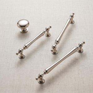 Può includere: Un set di ferramenta in nichel lucido che include un pomello rotondo per mobile e tre maniglie allungate per cassetto. Ogni maniglia presenta un design a barra sottile e affusolata con accenti sferici decorativi nei punti di fissaggio.