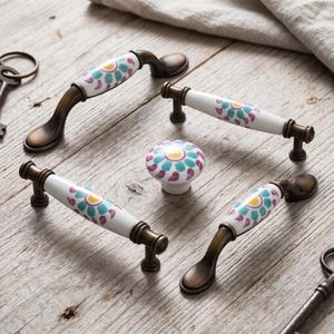 May include: A collection of four cabinet handles and one round knob featuring white ceramic centers with a floral pattern in teal, purple, and yellow. The hardware has an antique bronze finish with curved and straight metal mounting brackets.