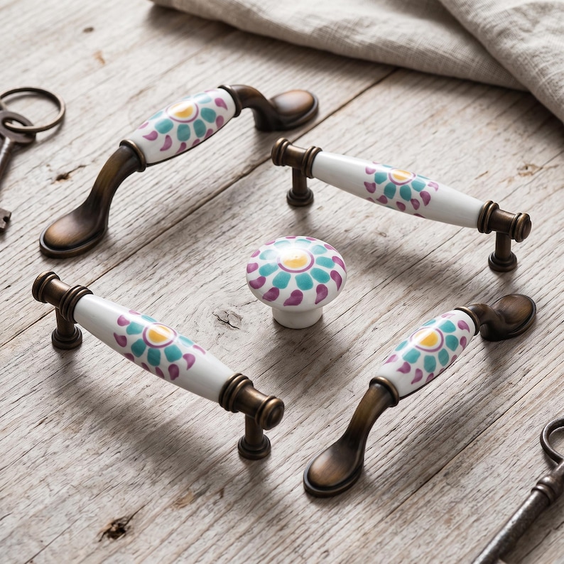 May include: A collection of four cabinet handles and one round knob featuring white ceramic centers with a floral pattern in teal, purple, and yellow. The hardware has an antique bronze finish with curved and straight metal mounting brackets.