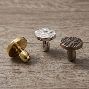 May include: Three mushroom-shaped cabinet knobs with round, textured tops and smooth, tapered stems. One knob is polished gold, one is polished silver, and one is a dark, brushed bronze finish, each featuring a unique, rippled surface pattern.