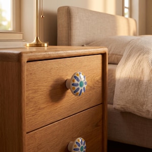 May include: A light oak wood nightstand featuring two drawers, each equipped with a round ceramic knob. The knobs are white with a hand-painted floral pattern consisting of blue petals, yellow dots, and a central teal circle, providing a decorative accent.