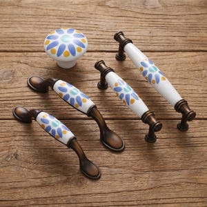 May include: A collection of cabinet hardware including one round ceramic knob and three arched metal drawer pulls. Each piece features a white ceramic base decorated with a repeating pattern of blue and yellow petals around a central light blue dot.