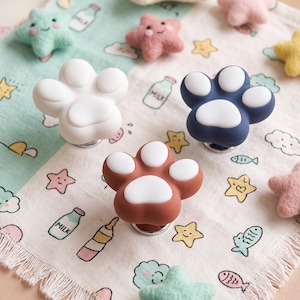 May include: Three paw-shaped cabinet knobs in white, dark blue, and terracotta, each featuring a white pad design. These functional hardware pieces are arranged on a patterned fabric surface decorated with small milk bottles, stars, and fish illustrations.