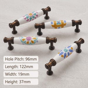 May include: Four ceramic drawer pulls with white bodies and bronze-toned metal ends. Each pull features a unique colorful floral or geometric pattern. Text indicates a hole pitch of 96mm, length of 122mm, width of 19mm, and height of 37mm.