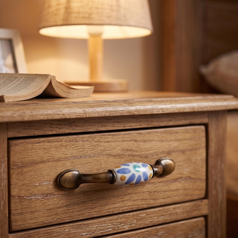 May include: A wooden nightstand drawer featuring a horizontal handle with a central ceramic insert. The ceramic piece is white with a hand-painted blue and yellow floral petal pattern, secured by aged brass-toned metal ends on the drawer front.