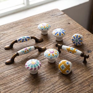 May include: A collection of ceramic cabinet hardware including two arched handles with antique brass finish and five round knobs. The white ceramic pieces feature hand-painted floral and geometric patterns in blue, yellow, teal, and purple colors.