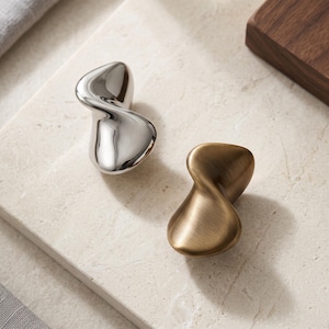 Peut inclure: Deux boutons de placard avec une forme organique, torsadée et abstraite. Un bouton a une finition argentée polie et réfléchissante, tandis que l'autre présente une finition dorée mate brossée. Les deux pièces ont un design incurvé lisse.