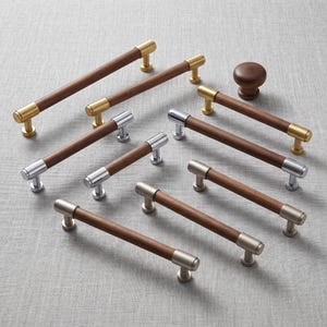 May include: A collection of cabinet hardware including several horizontal bar pulls with dark brown wooden centers and metallic end caps in brass, chrome, and brushed nickel finishes, alongside a single round dark brown wooden cabinet knob.