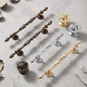 May include: A collection of metal cabinet hardware including three elongated drawer pulls and three round knobs. The hardware features ornate, textured detailing in antique brass, polished chrome, and brushed gold finishes with pointed ends.