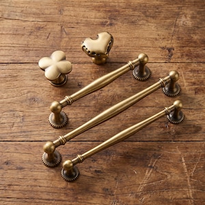 May include: A collection of antique brass hardware including two long drawer pulls with rounded ends, one clover-shaped knob, and one heart-shaped knob. Each piece features a smooth, metallic finish with a warm, golden-brown tone and circular bases.