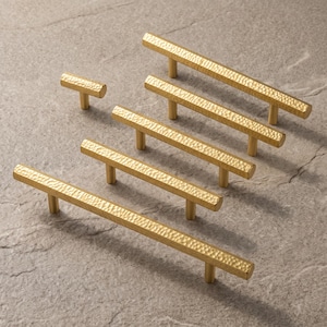 May include: A collection of gold-colored metal cabinet hardware including one small T-shaped knob and four elongated bar pulls. Each piece features a distinct hammered, textured surface finish and a straight, linear design with cylindrical posts.