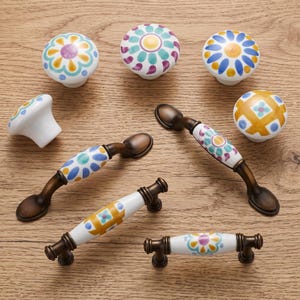 May include: A collection of ceramic cabinet hardware including four round knobs and three arched drawer pulls. The pieces feature white surfaces with hand-painted floral and geometric patterns in yellow, blue, teal, and purple, paired with bronze metal.