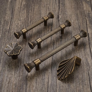 May include: A collection of antique brass hardware including three fluted bar pulls, one flower-shaped knob, and one fan-shaped pull. Each piece features a dark, weathered metallic finish with visible ridges and textured detailing on the surfaces.