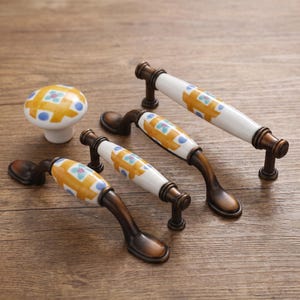 May include: A collection of cabinet hardware including one round knob and three arched drawer pulls. Each piece features a white ceramic center with a yellow grid pattern, blue dots, and small teal flowers, accented by dark bronze metal ends.