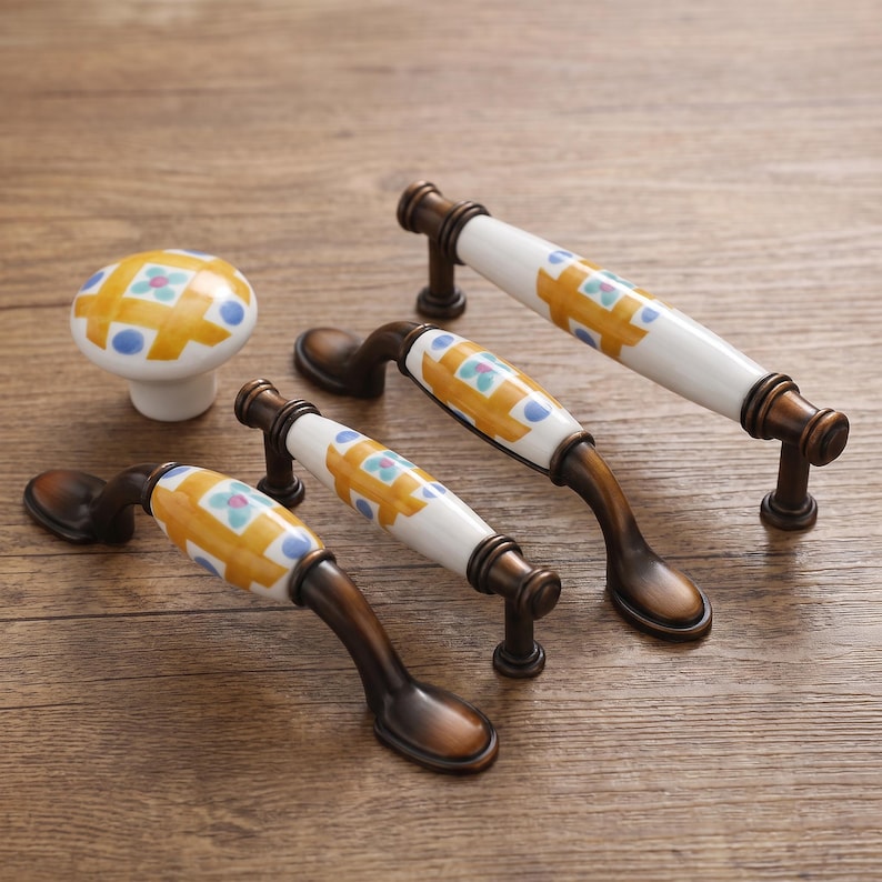May include: A collection of cabinet hardware including one round knob and three arched drawer pulls. Each piece features a white ceramic center with a yellow grid pattern, blue dots, and small teal flowers, accented by dark bronze metal ends.