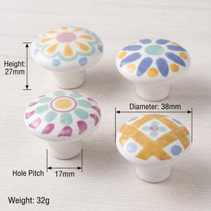 May include: Four round ceramic knobs with hand-painted floral and geometric patterns in yellow, blue, pink, and green. Each knob has a height of 27mm, a diameter of 38mm, a hole pitch of 17mm, and a weight of 32g, as indicated by the text labels.